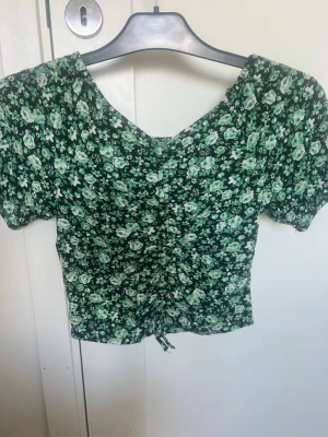 Grön croppad blus - Supersöt grön blommig blus med kort ärm och v-ringning. Toppen är rynkad framtill vilket ger en snygg och trendig effekt. Den är kort i modellen!!