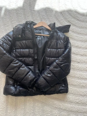 Svart pufferjacka från 157 - Snygg svart pufferjacka från 157 med huva och dragkedja framtill. Jackan är quiltad och har en glansig finish som ger en modern look. Luvan har en dragkedja som gör att den gör att ta av. Perfekt för kalla vinterdagar och enkel att matcha med olika outfits.