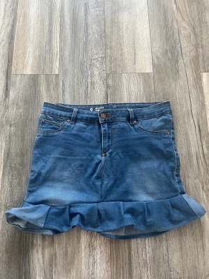 Blå jeanskjol med volangkant - Snygg blå jeanskjol från jegging jane med volangkant nertill. Kjolen har klassisk femficksdesign, bälteshällor och stängs med knapp och dragkedja. Perfekt för dig som gillar en trendig och ungdomlig stil.