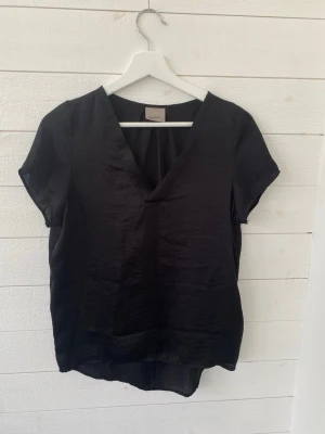 Svart blus med v-ringning - En stilren svart silkes blus med v-ringad hals och korta ärmar. Blusen har en något lös passform och rundad nederkant, vilket ger en avslappnad men ändå snygg look. Perfekt att matcha med jeans eller kjol för en trendig outfit.