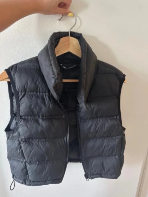 Svart dunväst med hög krage - Snygg svart dunväst med quiltad design och hög krage. Västen har en kortare passform och dragsko nertill för justerbar stil. Perfekt att slänga över en hoodie eller tröja för extra värme. Materialet är glansigt och lätt, vilket ger en modern look.