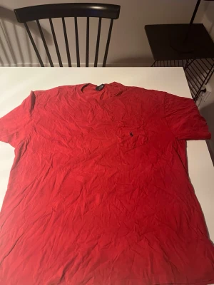 Röd t-shirt från Polo Ralph Lauren - Klassisk röd t-shirt från Polo Ralph Lauren med kort ärm och rund hals. Bröstficka med ikonisk broderad logga i mörkblått. Tillverkad i mjuk bomull för skön känsla. Perfekt till jeans eller shorts för en avslappnad look.
