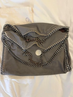 Grå Stella McCartney Falabella väska - Säljer en grå Falabella handväska från Stella McCartney med kedjedetaljer i silver runt kanterna och som axelrem. Väskan är i mjukt veganskt mockaliknande material och har ett ljusrosa innerfoder samt magnetlås. Perfekt för dig som gillar statement pieces. Den är köpt på NK i Stockholm för cirka 10 000kr. Den är i super bra skick men har två små fläckar på baksidan men inget som man ser när den används. Ett pytte litet hål på sidan men inte heller något som syns. Pris kan diskuteras!