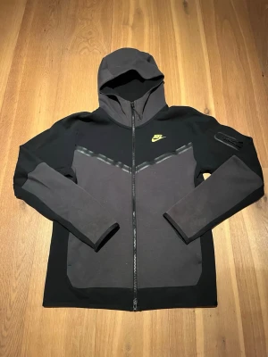 Nike Tech Fleece hoodie svart/grå - Snygg Nike Tech Fleece hoodie i svart och mörkgrått med dragkedja framtill och gul/grön Nike-logga på bröstet. Hoodien har huva, ficka på ärmen med dragkedja. Köpt på JD i London och knappt använt. Storlek S men funkar jättebra på en M också