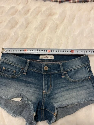 Hollister jeansshorts med slitningar - Snygga blå jeansshorts från Hollister med slitna detaljer och fransig kant. Klassisk femficksmodell med knapp och dragkedja framtill. Perfekta för sommaren och ger en avslappnad vibe. Gjorda i denim med cool tvätt och råa kanter.