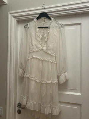 Vit volangklänning med öppen rygg - Romantisk vit klänning med volanger och transparenta långa ärmar. Djup v-ringning framtill och öppen rygg med dekorativa volangkanter. Kjolen är kort och har flera lager volanger. Materialet är tunt och skirt, troligen polyester eller liknande.