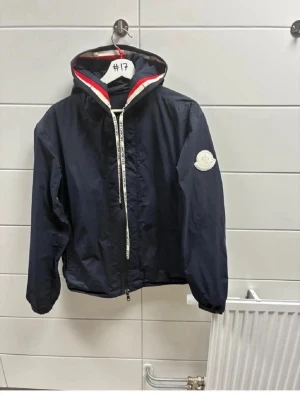Mörkblå äkta Moncler vindjacka med huva - Snygg mörkblå vindjacka från Moncler med huva och dragkedja framtill. Jackan har vita och röda detaljer på huvan samt Moncler-logga på ärmen. Perfekt för blåsiga dagar och enkel att matcha till olika outfits.