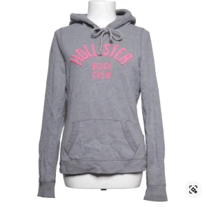 Grå hoodie från Hollister med tryck - Grå hoodie från Hollister med rosa tryck 'Hollister Beach Crew' på bröstet. Klassisk känguruficka framtill, dragsko i huvan och mjukt material.
