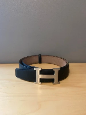 Svart H bälte - Stilrent svart bälte från med ikoniskt H-format spänne i silverfärg. Bältet har en klassisk design och är tillverkat i slätt skinn med beige insida. Perfekt accessoar för att lyfta vilken outfit som helst.