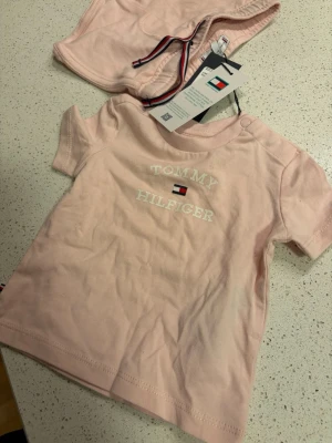 Rosa set från Tommy Hilfiger - Sött rosa set från Tommy Hilfiger med t-shirt och shorts. T-shirten har tryck med logga och text framtill, samt tryckknappar vid axeln. Shortsen har elastisk midja och dekorativt band i Hilfigers färger. Mjukt och bekvämt material, perfekt för varma dagar.