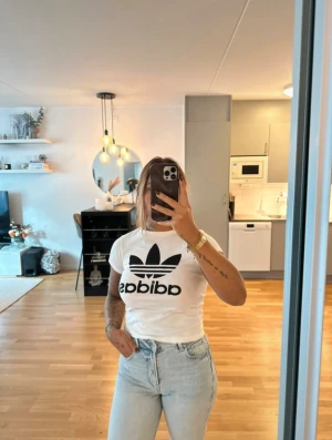 Vit Adidas t-shirt - Snygg vit t-shirt från Adidas med den klassiska svarta loggan tryckt framtill. T-shirten har korta ärmar, rund hals och är gjord i mjuk bomull. Perfekt till jeans eller shorts för en clean och sportig look. Jag är en s/m men passar även xs om man gillar att den sitter lite lösare