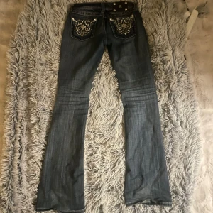 Mörkblå bootcut jeans med mönstrade fickor - Säljer ett par mörkblå bootcut jeans från Miss Me med snygga slitningar och broderade bakfickor med blommönster och nitar. Jeansen har låg midja och klassisk femficksdesign. Perfekta för dig som vill ha en unik och trendig look.innerben:81 cm, ytterben:101 cm, midja:37, benhöjd:17,5