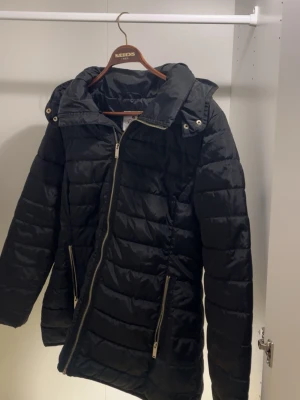 Svart lång dunjacka med huva - Snygg svart dunjacka med avtagbar huva och sneddraget blixtlås. Jackan har två dragkedjeförsedda fickor framtill och quiltad design som ger en modern look. Perfekt för kalla vinterdagar och riktigt varm.