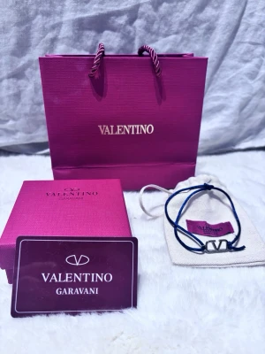 Valentino Garavani blå armband - Snyggt armband från Valentino Garavani med blå justerbar textilrem och en silverfärgad metallberlock i form av det ikoniska V-logotypen. Kommer med originalpåse, ask och presentpåse. Perfekt accessoar för dig som gillar exklusiva detaljer.