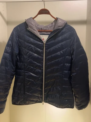 Mörkblå dunjacka med huva - Snygg mörkblå dunjacka med huva och dragkedja framtill. Jackan har ett quiltat mönster och grått innerfoder. Perfekt för kalla vinterdagar och har en modern, sportig look.