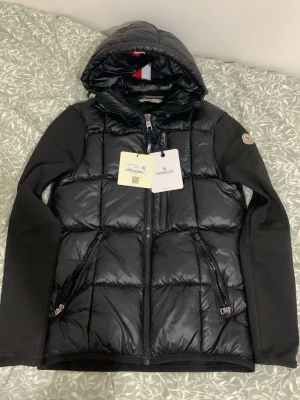 Svart pufferjacka med huva  från Moncler - Svart Cardigan från Moncler med glansig vadderad front, huva och ärmar i mjukt tyg. Jackan har två dragkedjefickor fram, Moncler-logga på ärmen och innerfoder med serietidningsmönster. Perfekt för kalla vinterdagar. Helt ny köpt från farfetch.com för ca 12,000kr. Skriv gärna om ni har frågor för och ställa!  200kr extra om du vill få med bägge påserna.