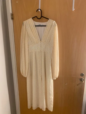 Ljusgul blommig långklänning från NA-KD - Söt långklänning i ljusgul ton med små blommor, v-ringad framtill och knappar längs hela framsidan. Klänningen har långa ärmar med lätt puff och smock i ryggen för skön passform. Storlek S och använd en gång. 