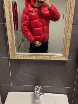 Röd pufferjacka från Polo Ralph Lauren - Säljer en röd pufferjacka från Polo Ralph Lauren med huva och dragkedja. Jackan har en blank finish, två sidofickor med dragkedja och en liten broderad logga på bröstet. Perfekt för kalla vinterdagar och riktigt snygg till streetwear-stilen.