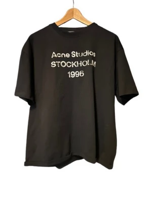 Svart Acne Studios t-shirt - Snygg svart t-shirt från Acne Studios med vit text på bröstet där det står 'Acne Studios STOCKHOLM 1996'. T-shirten har rund halsringning och korta ärmar. Perfekt för en clean och stilren look. Materialet känns mjukt och bekvämt.