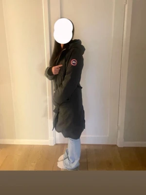Mörkgrön parkas från Canada Goose - Säljer en gröngrå jacka från Canada Goose med klassisk logga på ärmen. Jackan har huva, dragkedja och knappar framtill samt en längre modell som går ner över låren. Perfekt för kalla dagar och riktigt snygg till jeans.