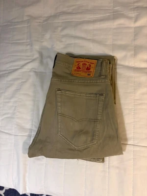Levis jeans  - Säljer ett par beige jeans från American Classic Jeans med rak passform och klassisk femficksdesign. Jeansen har en snygg patch bak i midjan och är tillverkade i ett slitstarkt bomullstyg. Perfekta för en avslappnad och stilren look.