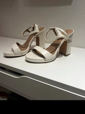 Vita chunky pumps med rem - Snygga vita pumps med bred klack och öppen tå. Skorna har en enkel rem över tårna och en justerbar ankelrem med silverdetalj. Tillverkade i ett slätt material som ger en stilren look. Perfekta för dig som gillar trendiga och bekväma klackar. Använda fåtal gånger och har kvar boxen de kom i🤗🤗🤗