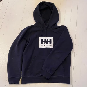 Hoodie Helly Hansen - Snygg marinblå hoodie från Helly Hansen med stor vit logga på bröstet. Tröjan är i väldigt gott skick. Storlek: Small