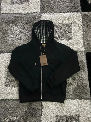 Svart Burberry hoodie med dragkedja - Snygg svart hoodie från Burberry med dragkedja framtill och klassiskt rutigt foder i huvan. Hoodien har justerbara snören vid huvan och ribbade muddar vid ärmar och nederkant. Perfekt för dig som gillar stilrena plagg med exklusiva detaljer.