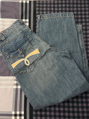 Blå jeans med broderade bakfickor - Snygga blå jeans med unika broderade detaljer i gult och vitt på bakfickorna. Klassisk femficksmodell med knapp och dragkedja. Materialet är klassisk denim.