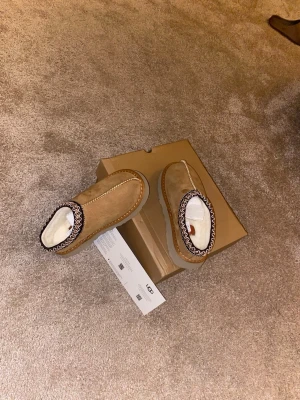 Bruna fårskinnstofflor från UGG - Mysiga bruna tofflor från UGG i mocka med vit fluffig fårskinnsinsida. Tofflorna har en dekorativ kant i svartvitt mönster runt öppningen och en rejäl sula i beige. Perfekta för att hålla fötterna varma hemma.