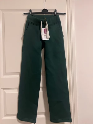 Russemerch Mörkgröna mjukisbyxor med raka ben - Mörkgröna mjukisbyxor med raka ben och bred resår i midjan samt vita dragsnören. Byxorna har två sidofickor och är tillverkade i ett mjukt, tjockt sweatshirtmaterial. Perfekta för en avslappnad och chill stil. Aldrig använt, beställt i fel storlek och glömt lämna tillbaka. 