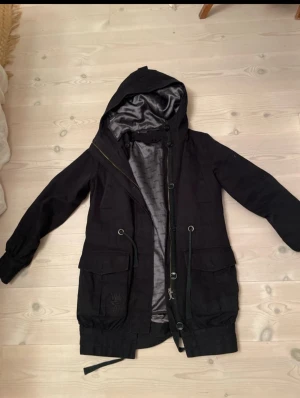 Svart parkasjacka med huva från WESC - Snygg svart parkasjacka med stor huva och dragsko i midjan. Jackan har två stora fickor framtill, knappar och dragkedja samt ett mjukt, glansigt innerfoder. Perfekt för kyliga dagar och enkel att styla till olika outfits. Löstagbart foder för en tunnare vårjacka. 