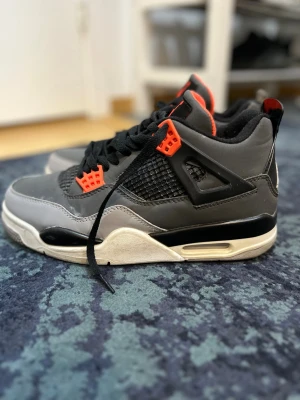 Jordan 4 Retro grå/svart/orange - Säljer ett par Jordan 4 Retro sneakers i grått, svart och vitt med orange detaljer. Skorna har meshpaneler på sidorna, svarta snören och den klassiska Jumpman-loggan på hälen. Ovandelen är i läder och syntetmaterial, och sulan är vit med svart och synlig Air-enhet.