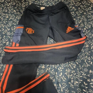 Manchester United träningsbyxor Adidas - Mörkblå träningsbyxor från Adidas med Manchester United-logga på ena benet och Adidas-logga på andra. Byxorna har röda ränder längs sidorna och dragkedjor vid bensluten. Perfekta för fotbollsträning eller gymmet.