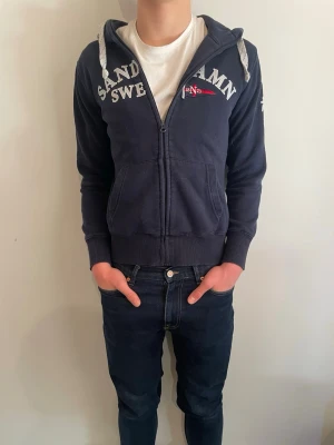 Marinblå hoodie Sandhamn - Marinblå hoodie från Sandhamn med dragkedja, vita tryck och broderade detaljer på bröstet och ärmen. Huva med vita snören och två fickor framtill. Perfekt för en avslappnad och sportig stil.