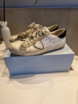 Phillipe model vita sneakers - Säljer ett par vita sneakers från Phillipe model med svarta detaljer på hälen och gråa inslag på sidorna. Skorna har snörning och rund tå, tillverkade i skinn och mocka. Perfekta för dig som gillar stilrena och lyxiga sneakers med cool design. Ja jag säljer dem för det priset