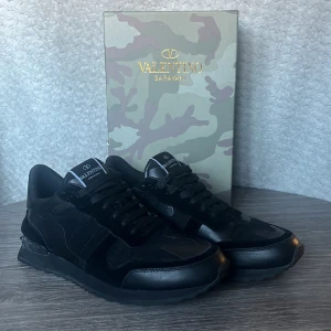 Svarta sneakers från Valentino Garavani - Sparsamt använda svarta sneakers från Valentino Garavani med kamouflagemönster och detaljer i både mocka och läder. Skorna har svart sula, svarta skosnören och logga på plösen. Perfekta nu i vinter och för dig som vill ha en exklusiv och trendig look.