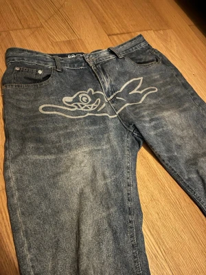 Jeans från Billionaire Boys Club - Unika blå jeans från Billionaire Boys Club med stort grafiskt tryck av en tecknad krokodil på framsidan och isglass-dödskalle på bakfickorna. Jeansen har en avslappnad passform, klassisk femficksdesign och är gjorda i slitstark denim. Perfekta för dig som vill sticka ut med din stil. Sitter baggy storlek L / ungefär 34