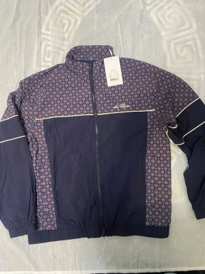 Mönstrad vindjacka från Les Deux - Snygg vindjacka från Les Deux i marinblått med färgglatt geometriskt mönster i rött, blått och vitt på axlar och ärmar. Jackan har hel dragkedja, hög krage och vita detaljer. Perfekt för dig som vill sticka ut med stilren design.