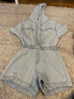 Ljusblå jeansjumpsuit med huva Zara - Supersnygg ljusblå jeansjumpsuit från Zara med korta ärmar, huva och knappar framtill. Resår i midjan ger en skön passform och shortslängd gör den perfekt för varma dagar. Tillverkad i denim för en cool och avslappnad look.