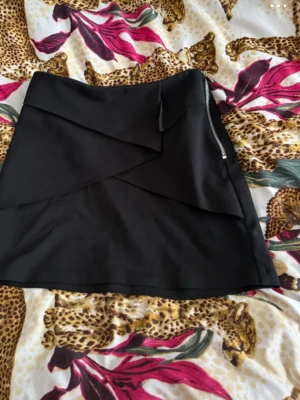 Svart omlottkjol med dragkedja - Snygg svart kortkjol med omlottdetaljer och dragkedja i sidan. Kjolen har en stilren look och passar perfekt till olika outfits. 