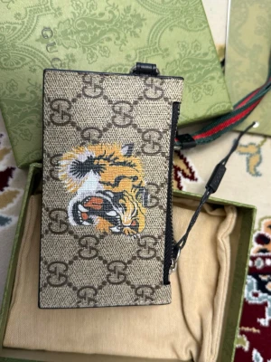 Gucci plånbok med tigertryck - Snygg plånbok från Gucci i beige canvas med klassiskt GG-mönster och färgglatt tigertryck på framsidan. Svarta läderdetaljer och flera kortfack på insidan. Kommer med avtagbar handledsrem och originalförpackning.