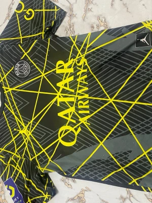 PSG Mbappé matchtröja svart/gul - Snygg PSG matchtröja i svart med gula grafiska linjer och detaljer. Framsidan har Qatar Airways-logga och PSG-emblem, baksidan har Mbappé och nummer 7 i gult. Tillverkad i lätt och ventilerande material, perfekt för fotboll eller träning. Passar ungefär 14 åringar, har lite trådar som sticker ut men inga problem alls.