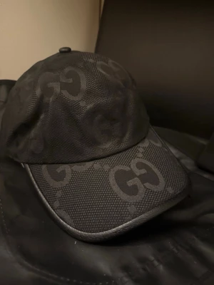 Svart GG-keps från Gucci - Säljer en svart keps från Gucci med det klassiska GG-mönstret över hela kepsen. Kepsen har böjd skärm och justerbart band bak i svart läder med guldfärgad detalj. Materialet är en mix av textil och läder, vilket ger en lyxig känsla och stilren look.