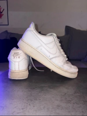 Vita Nike Air Force 1 sneakers - Klassiska vita Nike Air Force 1 sneakers i läder med vit sula och vita skosnören. Ikonisk Nike Swoosh på sidan och AIR-detalj på hälen. Låg modell som passar till det mesta och har perforeringar på tån för ventilation. Har några skavanker men syns på bilderna 