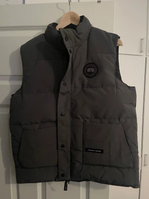 Mörkgrå dunväst från Canada Goose - Snygg mörkgrå dunväst från Canada Goose med hög krage, två stora fickor framtill och diskret logotyp på bröstet. Västen har dragkedja och tryckknappar samt innerficka. Perfekt för lager-på-lager och håller dig varm under kyliga dagar.