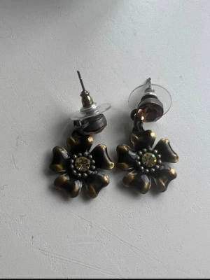 Blommiga örhängen i brons och guld - Unika örhängen i form av blommor med antik bronsfärg och guldfärgade detaljer. Blommorna har en glittrande sten i mitten och små pärlliknande dekorationer runt om. Örhängena har stift och rund, glittrig sten vid fästet. Perfekta för dig som gillar vintagekänsla. 👜💕