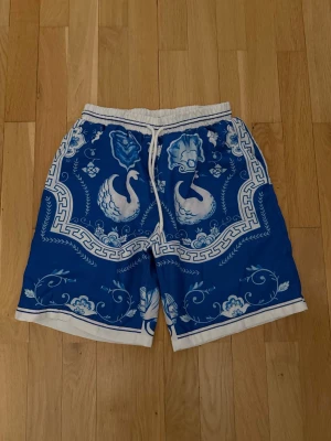 Casablanca shorts - Säljer ett par blåvita shorts med unikt mönster av svanar och blommor. Shortsen har elastisk midja med vit snörning och vita detaljer längs kanterna. Perfekta för sommaren och riktigt sköna att bära till vardags. L men passar typ M