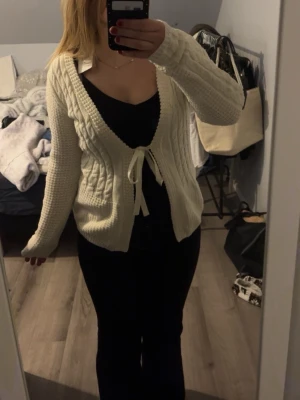 Vit stickad kofta med knytband - Mysig vit stickad kofta med långa ärmar och knytband framtill. Koftan har en djup v-ringning och ett grovt stickat mönster som ger en avslappnad och trendig look. Perfekt att slänga över en topp för en chill stil.