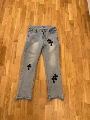 Chrome hearts jeans  - Unika ljusblå jeans med raka ben och råa kanter nertill. Dekorerade med flera svarta korsapplikationer både fram och bak. Jeansen har klassisk femficksdesign och är tillverkade i denim. Perfekta för dig som vill sticka ut med en edgy streetwear-look. Knappen är utbytt mot en annan från förra ägaren.